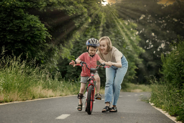 Mother teaching son to ride bicycle. Happy cute boy in helmet learn to riding a bike on the bike path in summer raining day at sunset time. Family weekend. - Foto, immagini