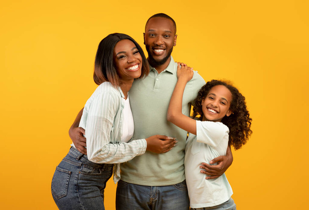 幸せなアフリカ系アメリカ人の母、父と彼らの小さな娘の肖像画黄色の背景の上にポーズ - 写真・画像