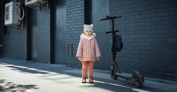 Carino piccola ragazza caucasica accanto allo scooter elettrico contro il muro di mattoni scuri - Foto, immagini