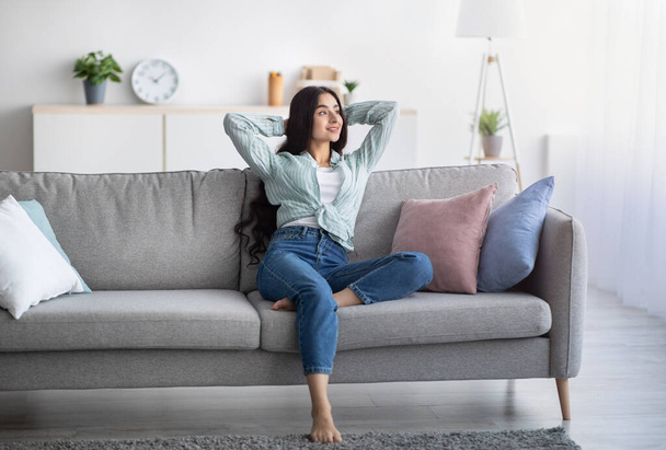 Täysi pituus nuori intialainen nainen rentouttava laiska viikonloppuna kotona, istuu sohvalla kädet pään takana, kopioi tilaa - Valokuva, kuva