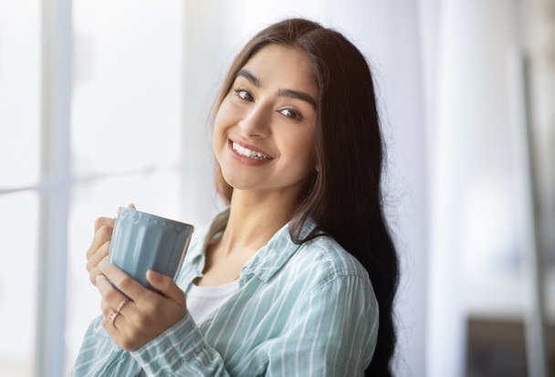Portrait of pretty Indian lady standing near window with cup of hot drink, smiling at camera, indoors - Fotó, kép