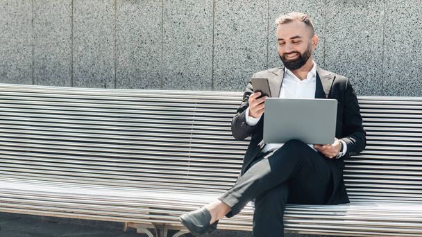 Зрелый бизнесмен в костюме с ноутбуком сидит на скамейке и использует смартфон на открытом воздухе, панорама с бесплатным пространством - Фото, изображение