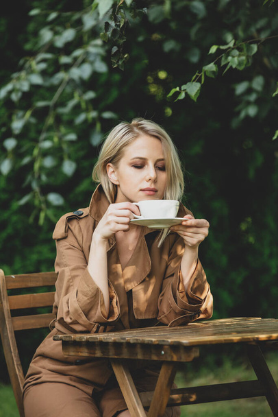 Schöne Frau im Mantel mit Tasse Kaffee am Holztisch in einem Garten mit Bäumen im Hintergrund - Foto, Bild