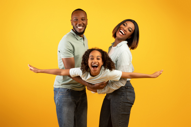 Koncepcja rodzinnej więzi. Radosny czarny mężczyzna i kobieta trzymając córkę na rękach, bawiąc się nad żółtą ścianą studio - Zdjęcie, obraz