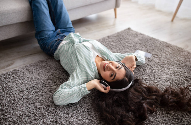 Acima vista da adorável senhora indiana em fones de ouvido ouvindo música, deitado no tapete macio em casa - Foto, Imagem