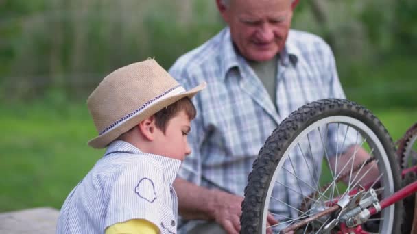 allegro bambino maschio, insieme al nonno premuroso, ripara la ruota della bicicletta mentre cammina fuori città - Filmati, video