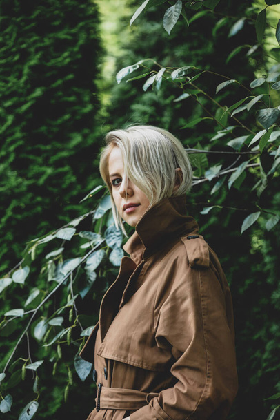 Style femme en manteau aux cheveux blonds rester à côté des arbres dans un jardin  - Photo, image
