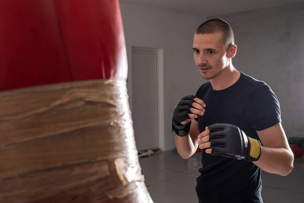 Молодой человек Мма спортсмен тренировки бокса удары тяжелую сумку с перчатками на руках в спортзале академии боевых искусств реальные люди копируют пространство - Фото, изображение