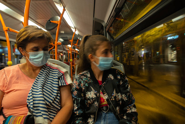 Middelbare leeftijd en jonge vrouw gezeten met masker door covid19 op een stadsbus als het reist door de stad concept vervoer met selectieve focus - Foto, afbeelding