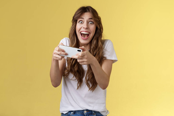 Extremely happy thrilled playful girl gamer playing awesome great new smartphone game hold mobile phone horizontal cheering look camera astonished impressed beating record yellow background - Photo, Image