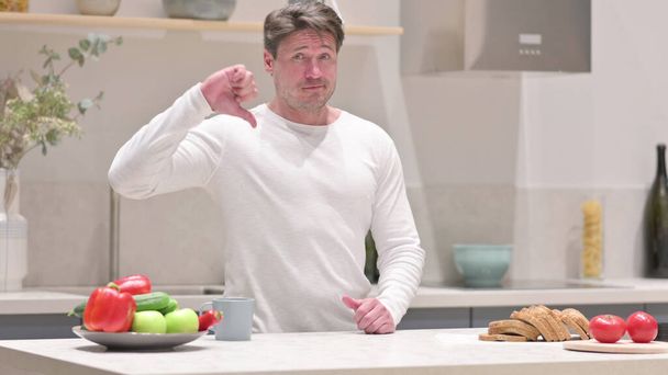 Middle Aged Man Showing Thumbs Down while Standing in Kitchen - Фото, изображение