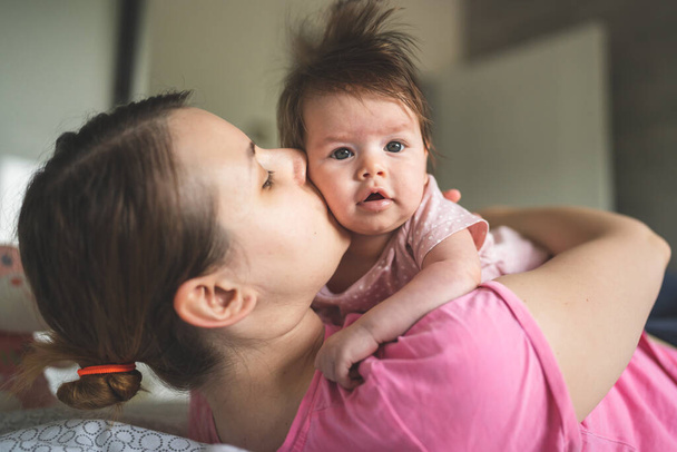 Young Adult caucasian mother playing with her four months old baby daughter while lying and kissing on the bed in bedroom at home parenting and motherhood family and happiness love concept - Photo, Image