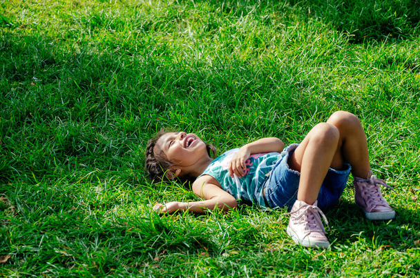 A little girl is smiling lying on the bright green grass. Portrait of a happy child on the grass in summer. The view from the top. Space for text. - 写真・画像