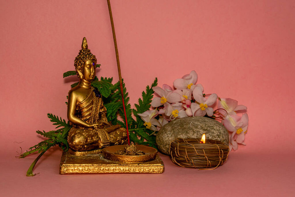 A closeup shot of a small statue with a candle and flowers on a pink background - Фото, изображение