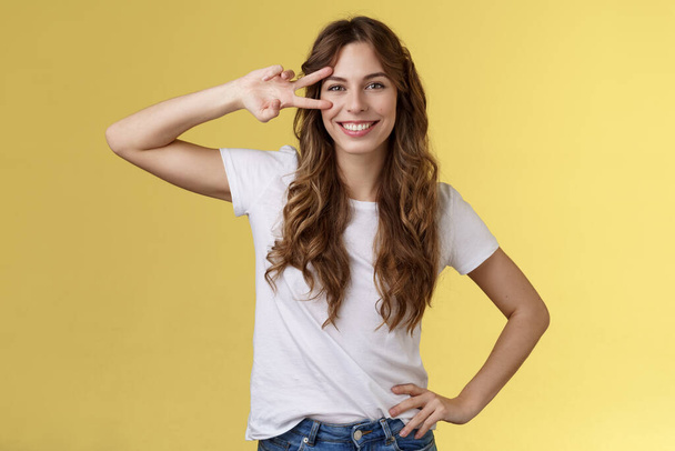 Cheerful positive cute girl curly-haired show peace sign delighted enjoy youth having perfect summer holidays determined rest trip vacation smiling broadly stand confident yellow background - Photo, Image