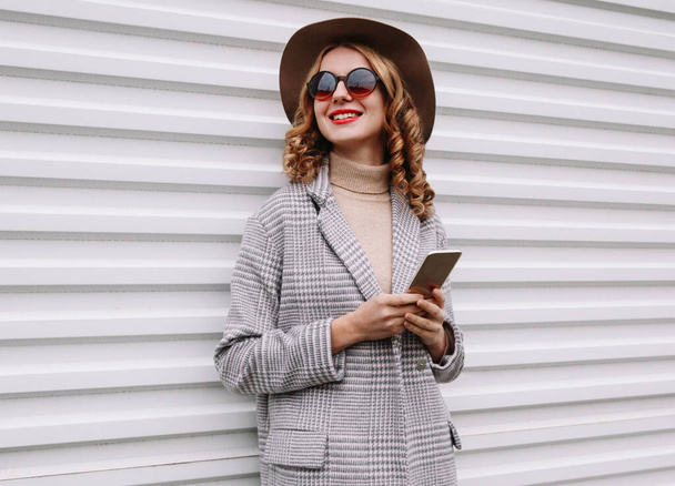 Ritratto di giovane donna sorridente elegante con smartphone che distoglie lo sguardo indossando un cappotto a scacchi, cappello rotondo su sfondo bianco - Foto, immagini
