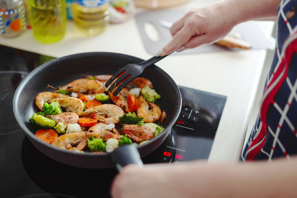 Woman staying at home kitchen and cooking shrimps with vegetables on pan. Home cooking or healthy cooking concept - Фото, изображение