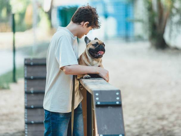 十代の若者は犬のための特別な遊び場で彼のかわいいフランスのブルドッグを教えています。少年は公園で赤いブルドッグとコマンドを学ぶ。犬を外で練習しなさい。子犬との友情はまだ生きてる - 写真・画像