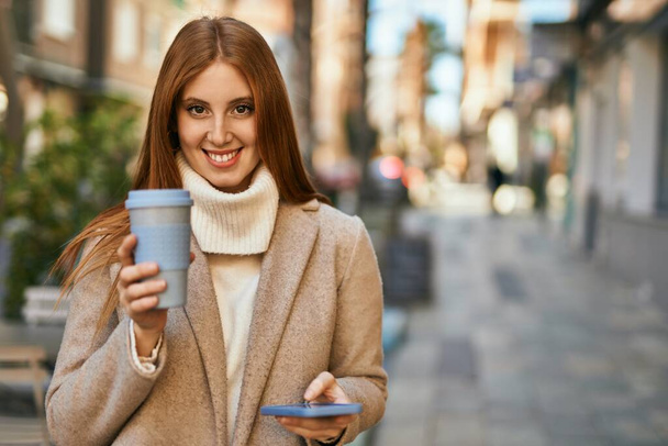 Junges rothaariges Mädchen trinkt mit Smartphone Kaffee in der Stadt. - Foto, Bild