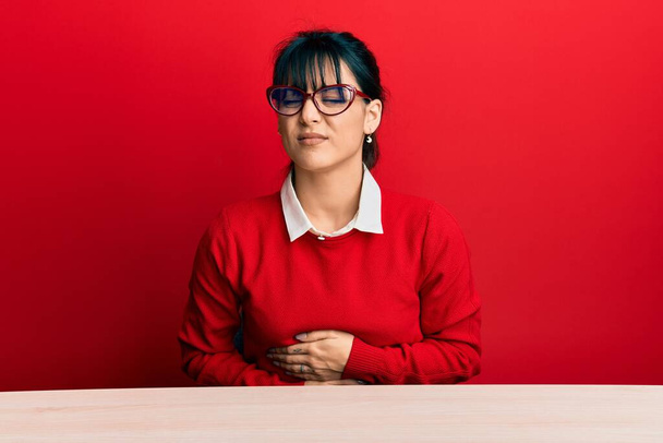 Young brunette woman with bangs wearing glasses sitting on the table with hand on stomach because nausea, painful disease feeling unwell. ache concept.  - Foto, imagen