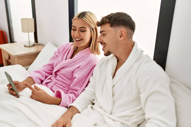 Joven pareja caucásica sonriendo feliz usando smartphone sentado en la cama en casa. - Foto, imagen