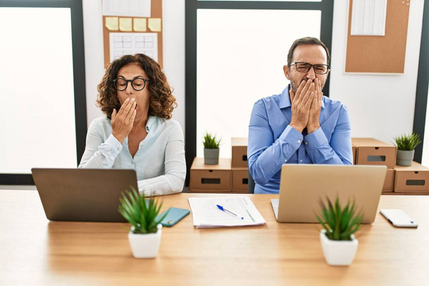Middle age hispanic woman and man sitting with laptop at the office bored yawning tired covering mouth with hand. restless and sleepiness.  - Photo, Image
