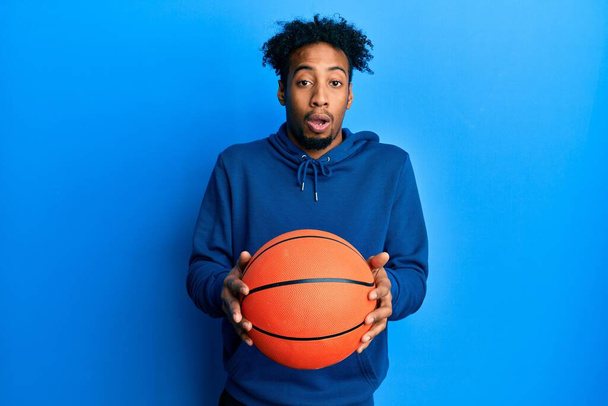 Young african american man with beard holding basketball ball afraid and shocked with surprise and amazed expression, fear and excited face.  - Photo, Image