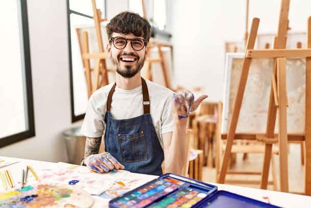 Spanischer Mann mit Bart im Kunststudio lächelt mit glücklichem Gesicht und zeigt mit erhobenem Daumen zur Seite.  - Foto, Bild
