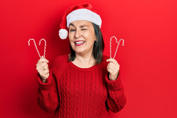 Mulher hispânica de meia idade usando chapéu de Natal segurando doces piscando olhando para a câmera com expressão sexy, rosto alegre e feliz.  - Foto, Imagem