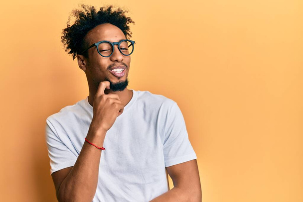 Νεαρός Αφροαμερικάνος με γενειάδα που φοράει λευκό πουκάμισο και γυαλιά που σκέφτεται συγκεντρωμένα την αμφιβολία με το δάχτυλο στο πηγούνι και αναρωτιέται  - Φωτογραφία, εικόνα