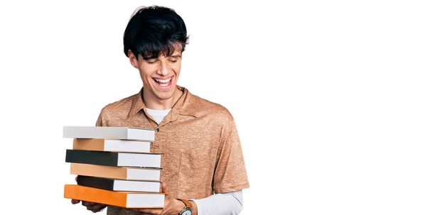 Handsome hipster young man holding a pile of books smiling and laughing hard out loud because funny crazy joke.  - Zdjęcie, obraz