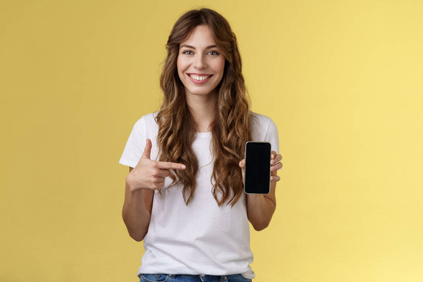 Girl have everything need smartphone. Cheerful relaxed carefree woman long curly hairstyle hold mobile phone pointing forefinger cellphone smiling broadly explain how app works yellow background - Fotografie, Obrázek
