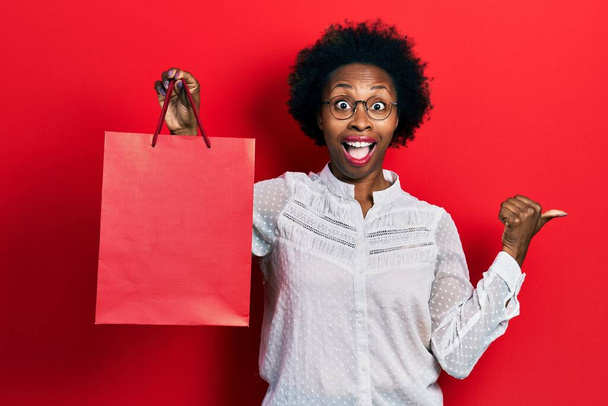 Young african american woman holding shopping bags pointing thumb up to the side smiling happy with open mouth  - Фото, зображення