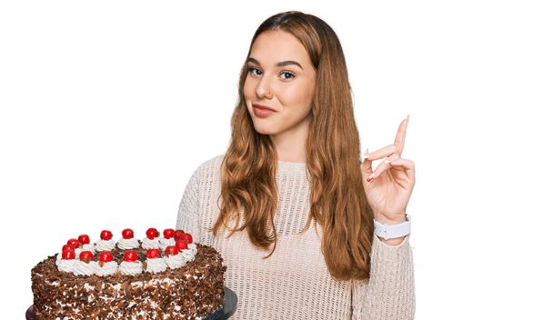 Joven mujer rubia celebrando cumpleaños sosteniendo gran pastel de chocolate sorprendido con una idea o pregunta apuntando con el dedo con la cara feliz, número uno  - Foto, imagen