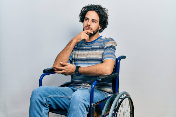 Hezký hispánec sedící na vozíčku s rukou na bradě a přemýšlející o otázce a zamyšleném výrazu. s zamyšleným úsměvem. pochybný koncept.  - Fotografie, Obrázek