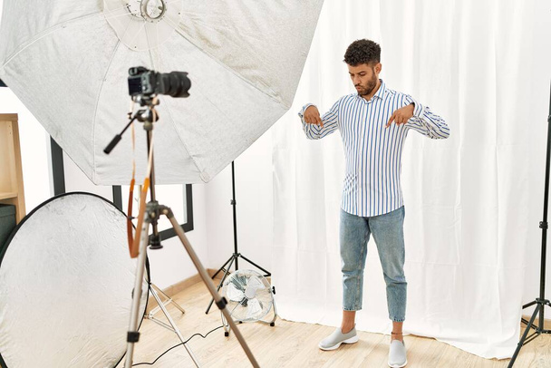 Arabo giovane uomo in posa come modello in studio di fotografia che punta verso il basso con le dita mostrando pubblicità, volto sorpreso e bocca aperta  - Foto, immagini