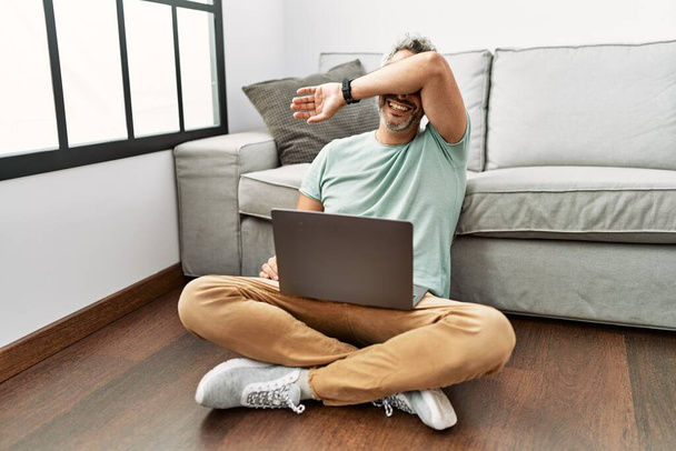Hispánec středního věku používá notebook, který sedí na podlaze v obývacím pokoji a vesele se usmívá, když si hraje s vystrčenýma rukama. překvapený a ukončený  - Fotografie, Obrázek