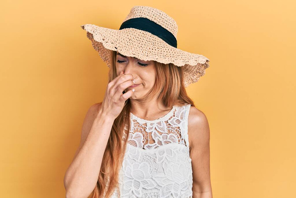 Mulher caucasiana jovem usando chapéu de verão cheirando algo fedorento e nojento, cheiro intolerável, segurando a respiração com os dedos no nariz. mau cheiro  - Foto, Imagem