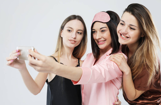 friendship, people, and technology concept - three young women friends wearing pajamas with smartphone taking selfie over grey background - Fotoğraf, Görsel