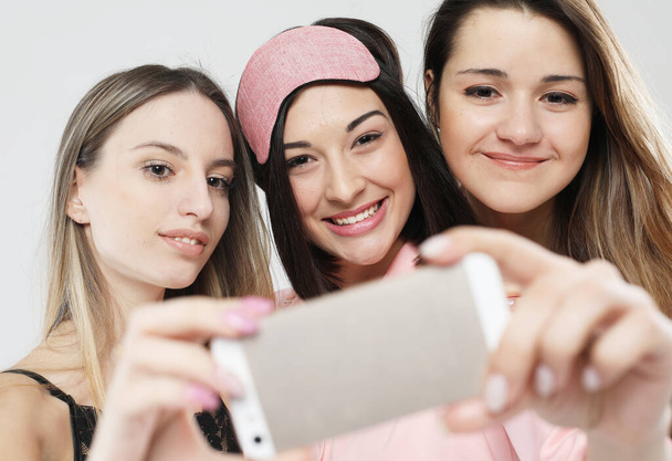 friendship, people, and technology concept - three young women friends wearing pajamas with smartphone taking selfie over grey background - Foto, immagini