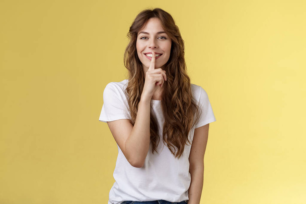 Cute cunning lovely european girl curly hairstyle hiding beauty secret smiling sensually show hush shush gesture index finger pressed lips grinning joyfully stand yellow background - Photo, Image
