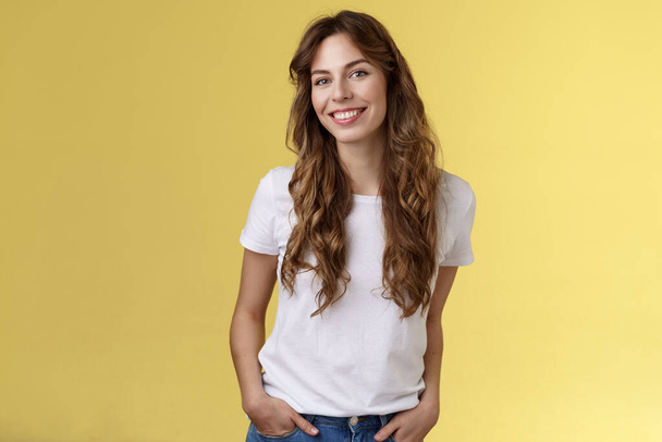 Attractive carefree confident friendly-looking charming feminine ubran woman long curly hair smiling toothy pleased tilt head intrigued hold hands pockets self-assured pose stand yellow background - Foto, Imagen