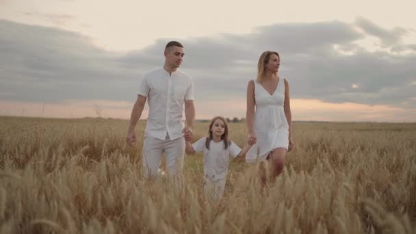Young couple of parents with girl children holding hands of each other and running through wheat field at sunset. Happy family jogging among barley meadow and enjoying nature together. Slow motion - Footage, Video