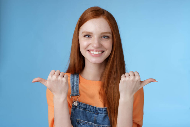 Indoor shot charismatic assertive happy smiling redhead woman orange shirt denim overalls pointing sideways thumbs left right showing choices opportunities give chance choose, blue background - Fotografie, Obrázek