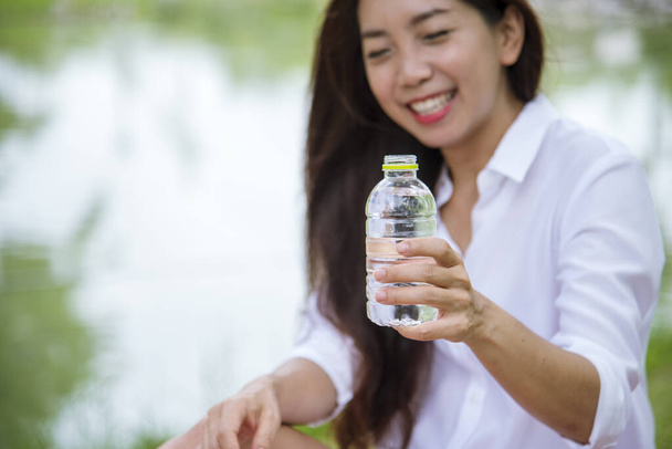 Щастя, прекрасні азійські гомілки, які тримають пляшку мінеральної води. Молода жінка п'є воду з пляшки в зеленому садовому парку. Азійка, яка п "є пляшку з водою, щаслива і посміхається - Фото, зображення