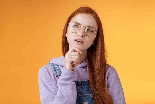 Thoughtful young creative smart redhead girl thinking figure out important thing standing upper left corner squinting thinking get clue touch chin thinking, pondering choice, orange background - Photo, Image