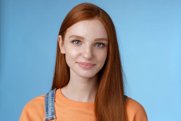 Выходящая молодая рыжая девушка голубые глаза в оранжевой футболке комбинезон улыбается приятно небрежно говорить стоя хорошее настроение радостные эмоции синий фон, слушать интересный разговор - Фото, изображение