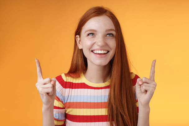 Счастливая веселая рыжая девушка веселится парк развлечений смеется радостно указывая вверх указательные пальцы смотреть вверх наслаждаться развлечением стоя оранжевый фон забавно улыбается радостно - Фото, изображение