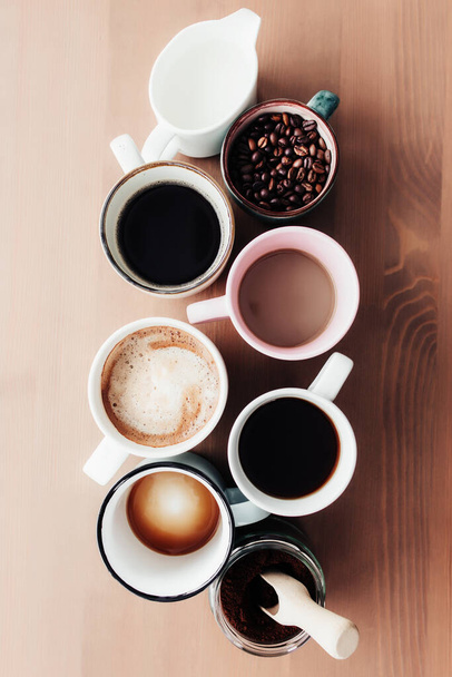 Mehrere Kaffeetassen und bunte Becher mit American, Espresso, Latte und Cappuccino, Milchglas, gerösteten Kaffeebohnen in Glasflasche, Holzlöffel und gemahlenem Kaffee in Glas auf Holzgrund - Foto, Bild