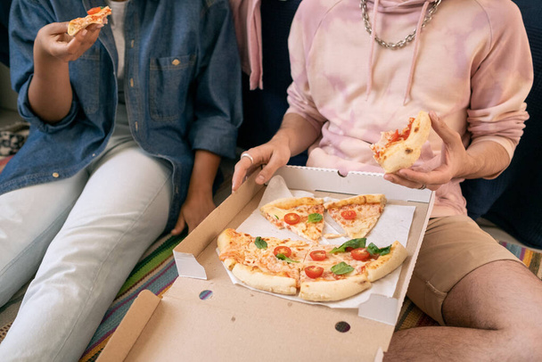 Двоє підлітків в повсякденному одязі їдять піцу, сидячи на підлозі
 - Фото, зображення